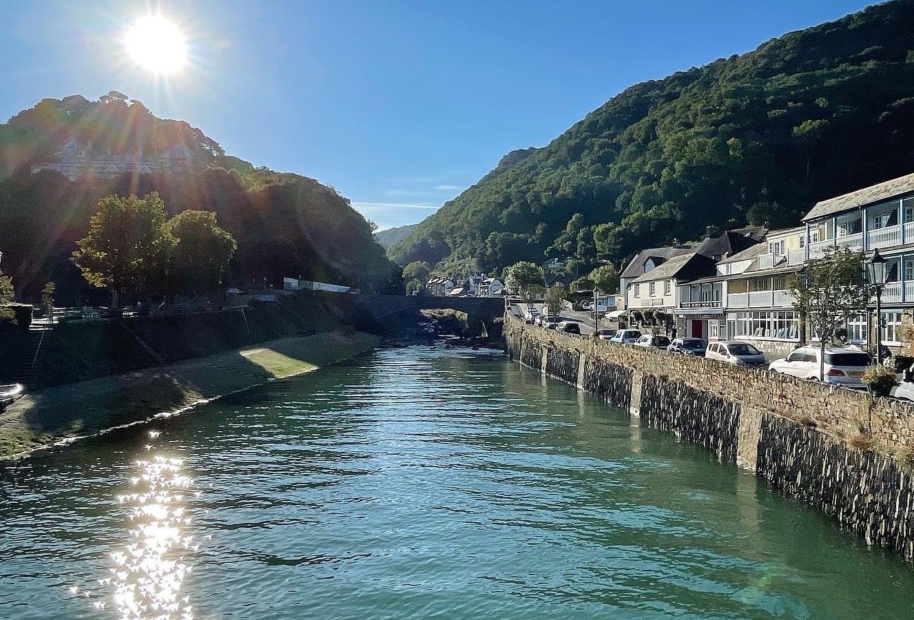 A serene view of a riverside town under a clear blue sky. The sun is shining brightly over green hills, casting reflections on the calm river. Houses and a few cars line the right side of the river, with a small stone bridge visible in the background—a perfect scene for Moor and Sea Holidays.