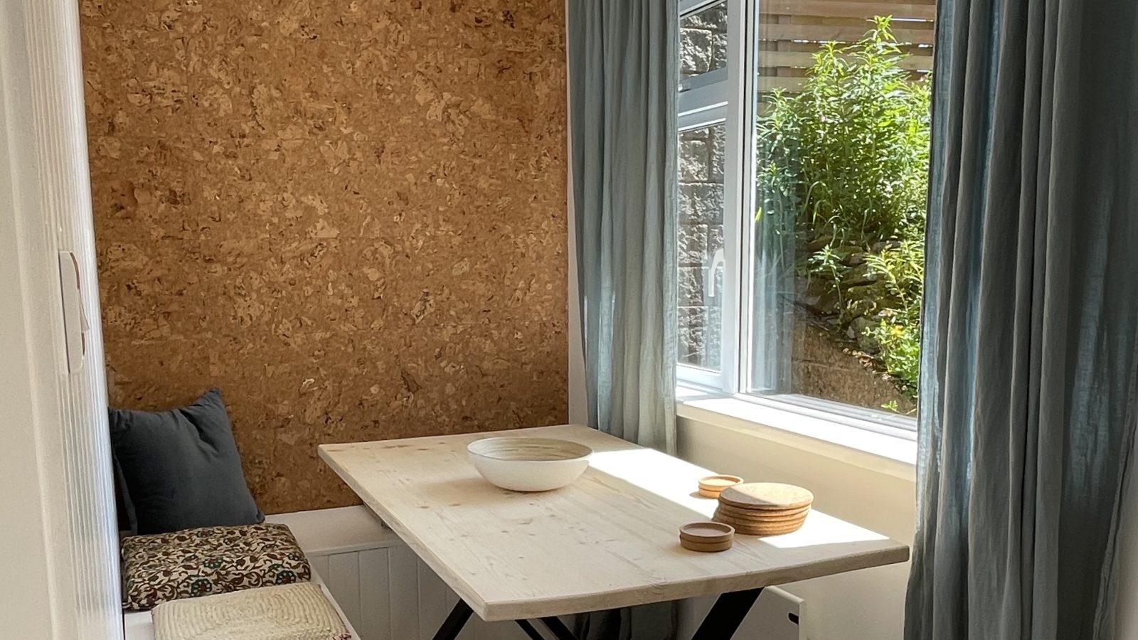 A cozy breakfast nook with a white wooden table and bench seating along one side, adorned with floral cushions. Next to the table, a large window with grey curtains lets in natural light. A wooden wall on the left adds rustic holiday charm, with a glass pendant light above.