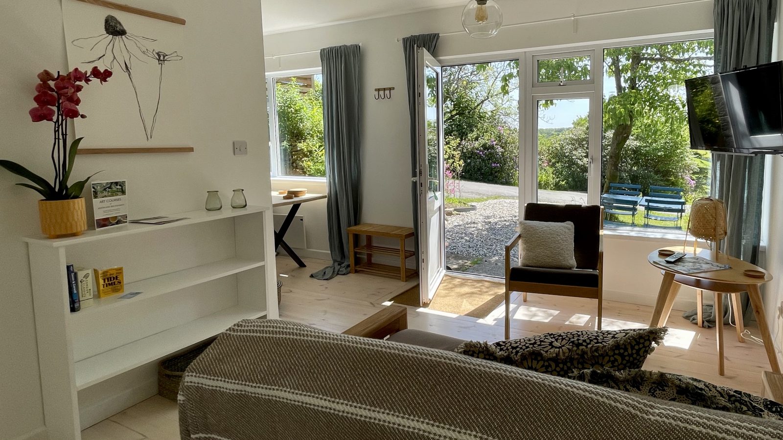 A bright living room featuring a brown chair, small round table, TV, and large windows with curtains overlooking a garden with trees and gravel path. A cozy sofa with a holiday blanket, a wall shelf with decorative items from seaside trips, and a desk are also visible.