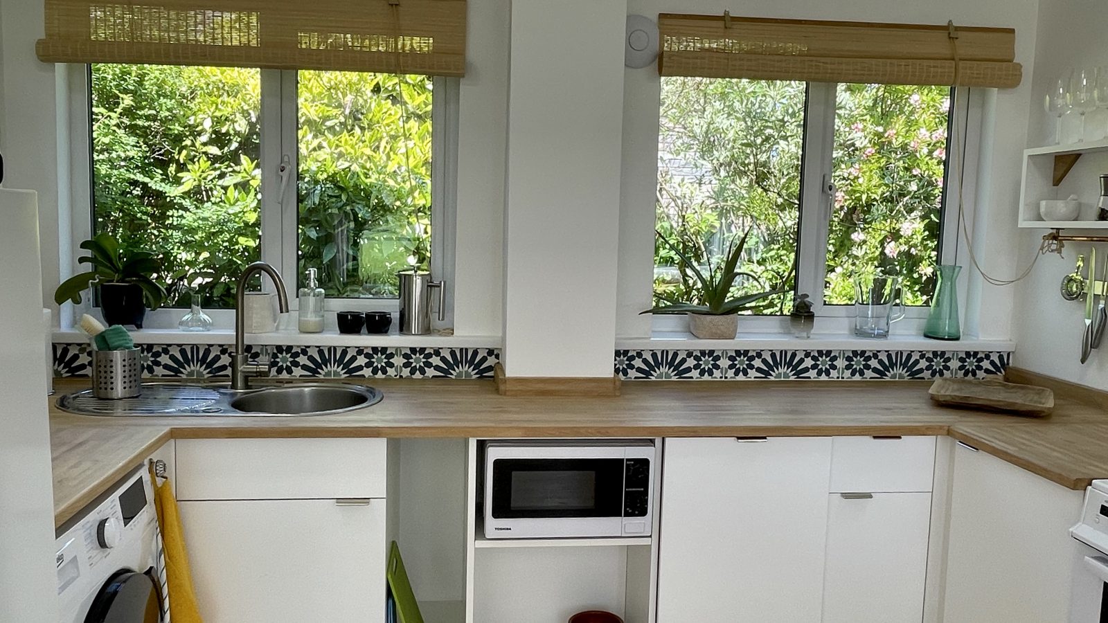 A modern kitchen with two windows showing a lush, green outdoor view evokes the tranquility of holiday getaways. The wooden countertops sit above white cabinets, while the backsplash features a black and white geometric pattern. A microwave is built into the cabinetry, and a washing machine is visible.