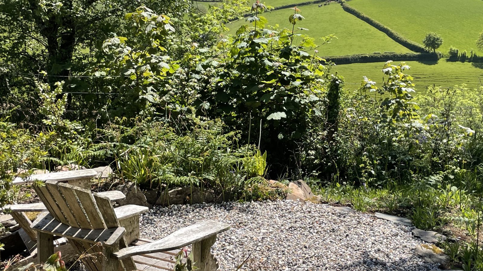 A peaceful outdoor seating area with two wooden chairs on a gravel surface, nestled among lush greenery. The scene overlooks rolling green hills and fields under a clear blue sky, offering a serene and picturesque view reminiscent of carefree holidays.