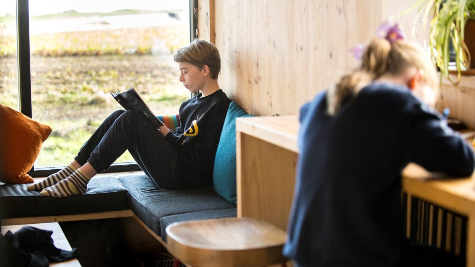 Two children are indoors at the serene Cambo Estate: one reads by a window where soft light filters in, and the other writes diligently at a desk, perhaps dreaming of future off-grid travels.