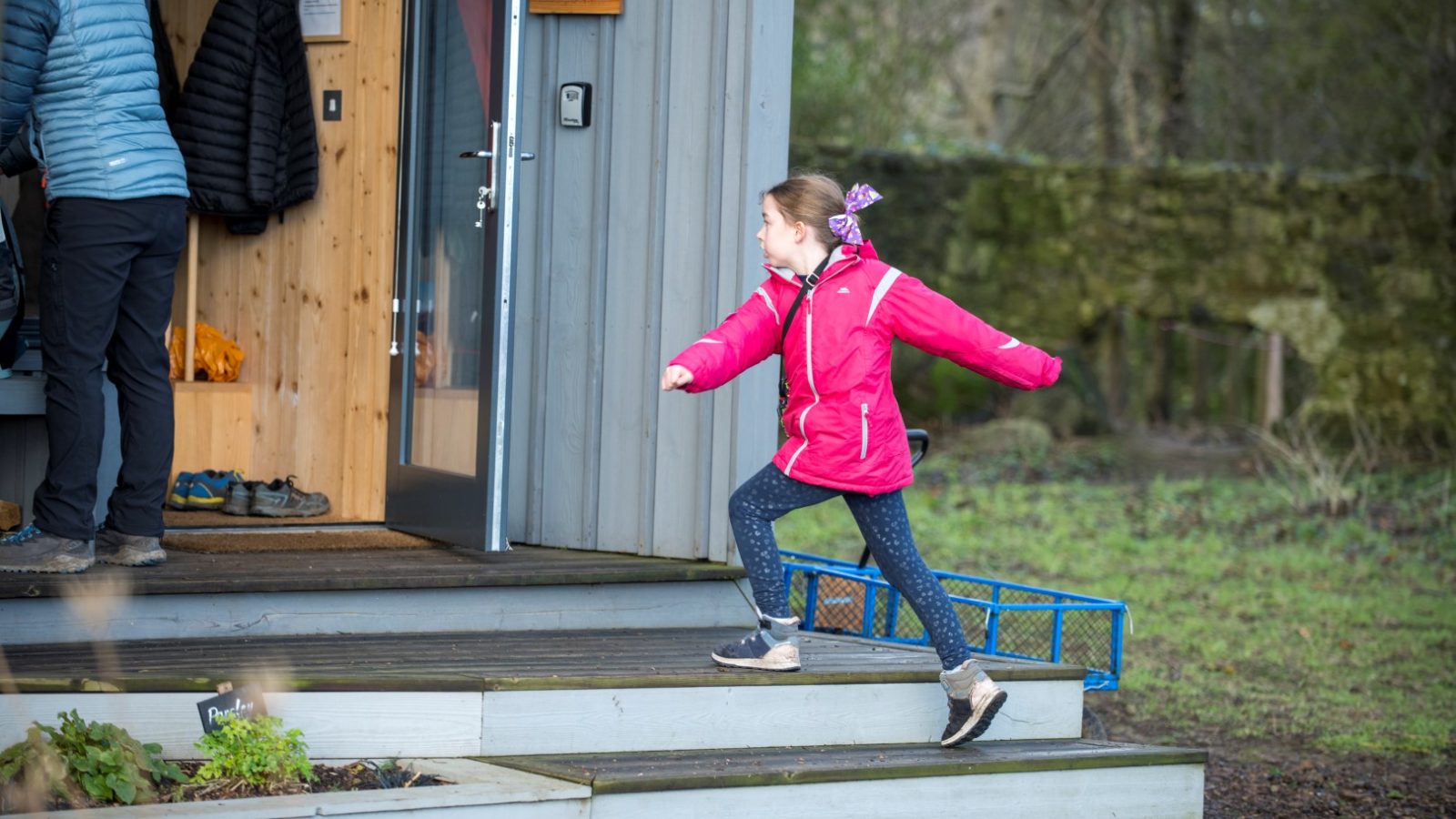 A girl in a pink jacket joyfully runs up wooden steps toward an open doorway on a cloudy day, embodying the spirit of off-grid travel at Cambo Estate.