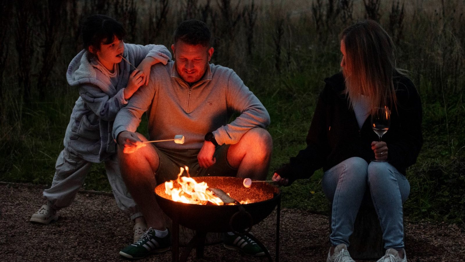 Three people relaxing around a fire pit, roasting marshmallows outdoors, with one holding a wine glass, at the serene Cambo Estate. Embrace the tranquility of off-grid travel in this idyllic evening setting.