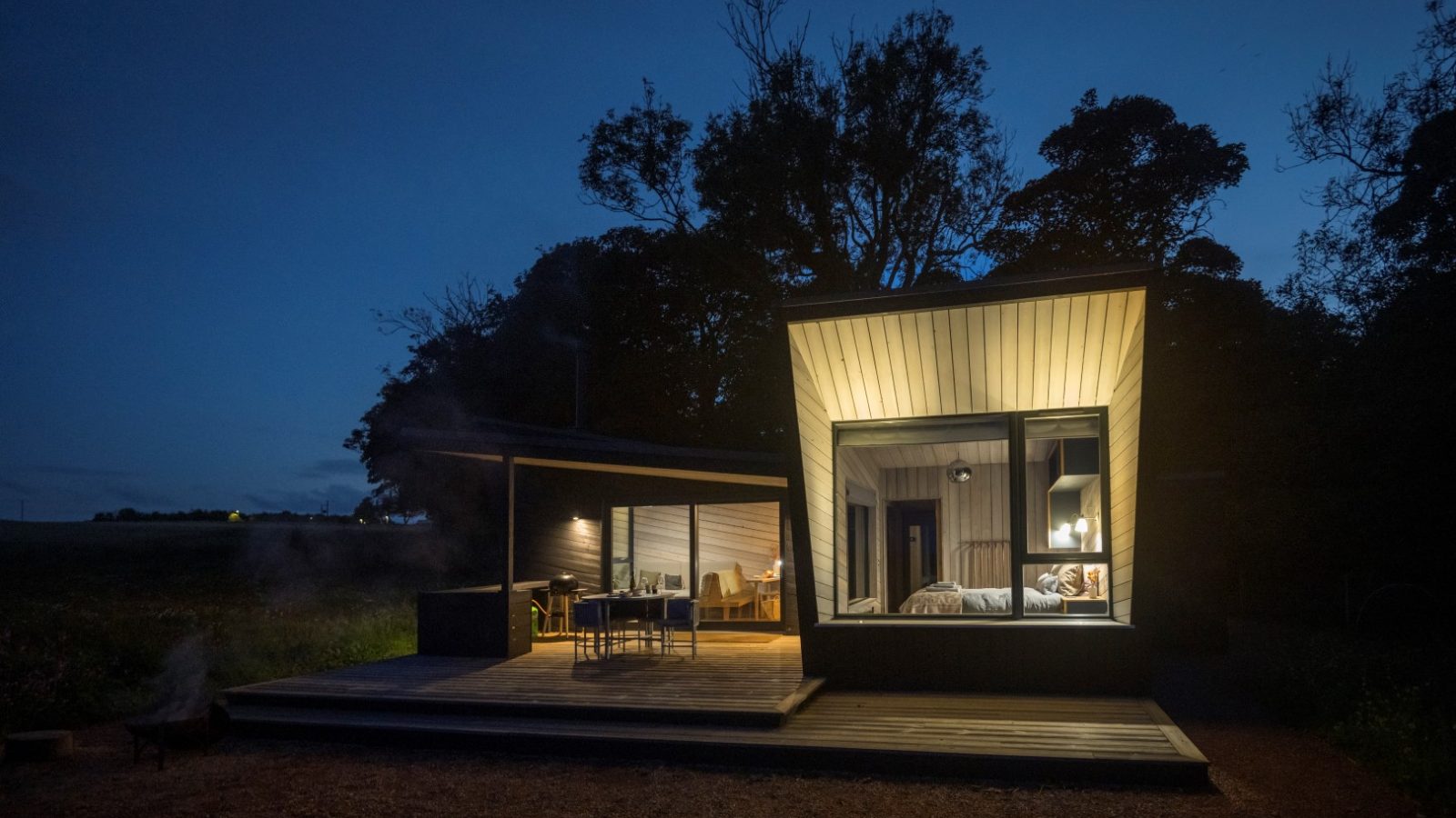 Nestled at Cambo Estate, this modern house features large illuminated windows and outdoor seating, surrounded by trees under a twilight sky. Perfect for those seeking off-grid travel with serene natural beauty.