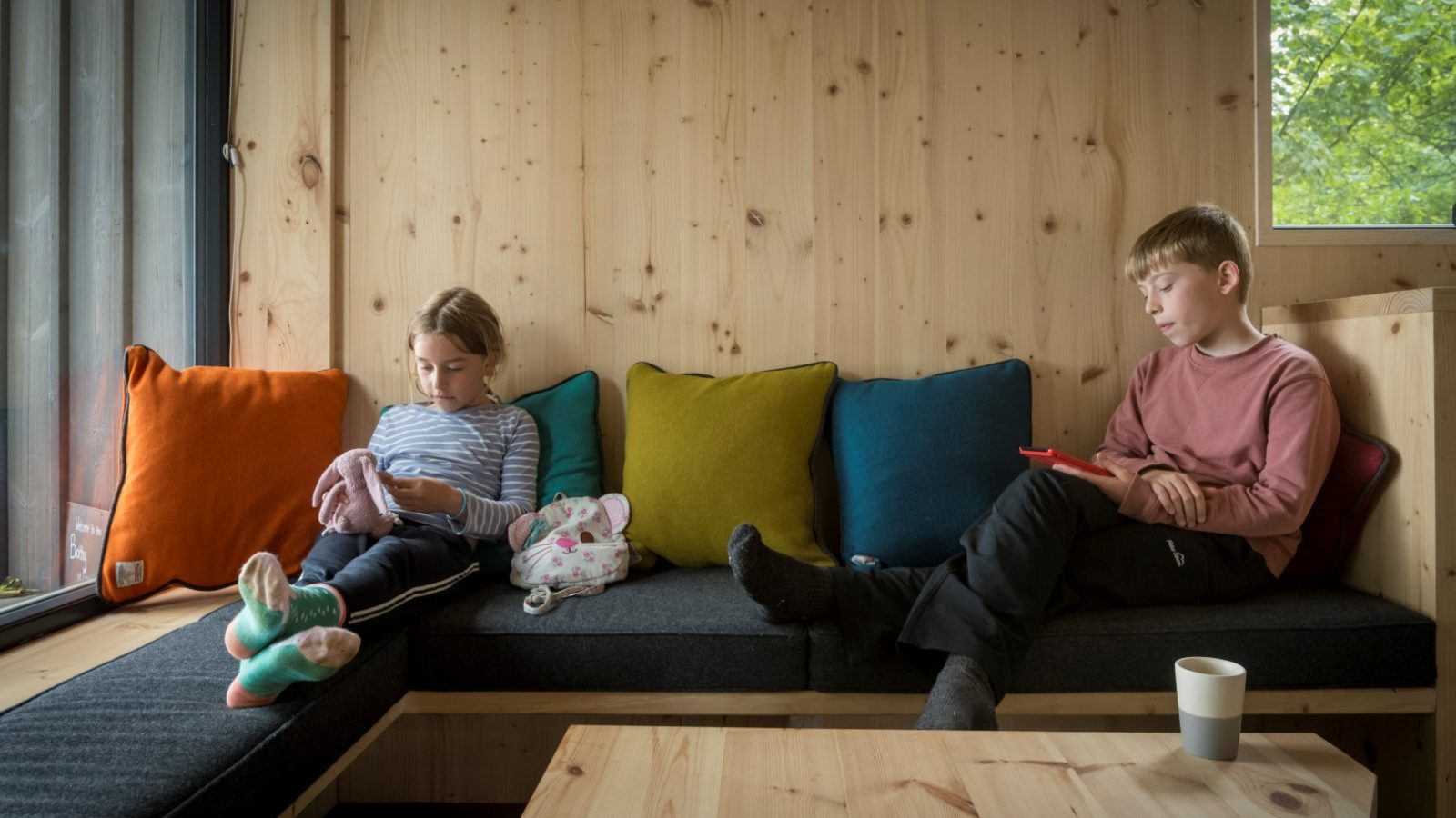 Two children sit on a wooden bench with colorful cushions at the tranquil Cambo Estate. Each is engrossed in their own activities, one with a toy and the other absorbed in a tablet, capturing the essence of serene off-grid travel.