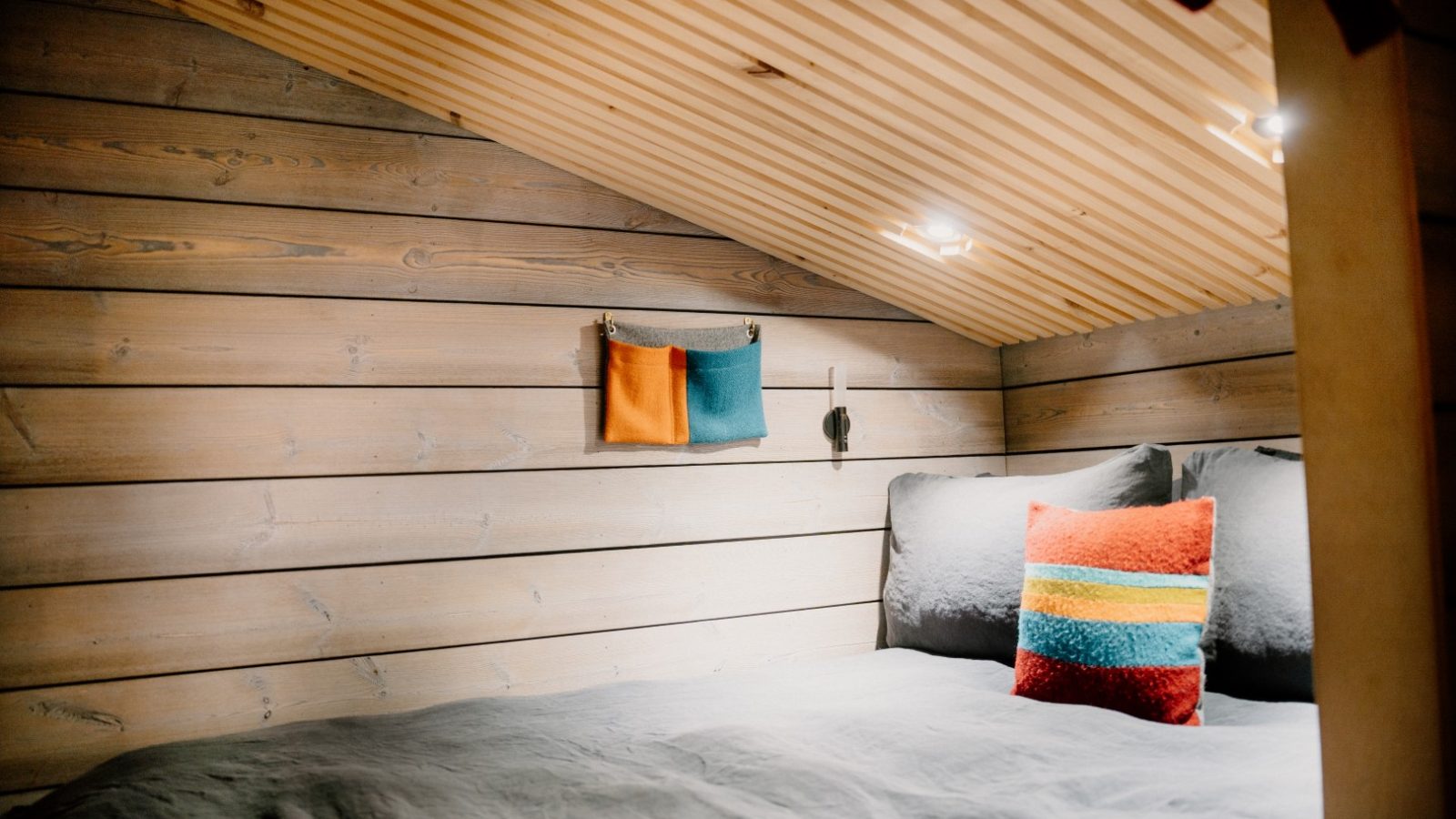 Cozy attic bedroom at Cambo Estate with a slanted wood ceiling, gray bedding, and a colorful pillow. The wall art features vibrant orange and teal fabric, perfect for your off-grid travel retreat.