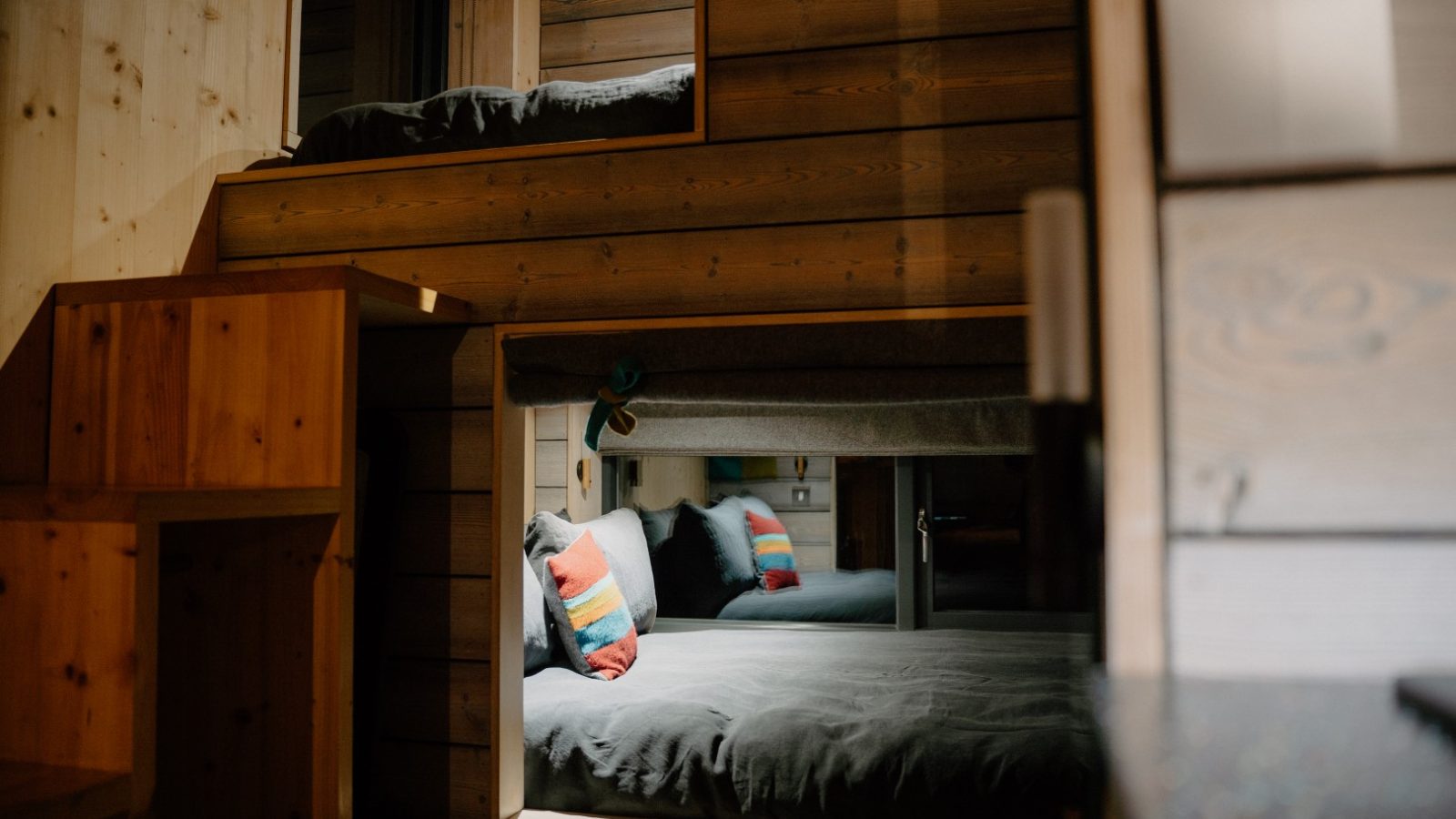 Bunk beds with colorful pillows in a modern wooden interior, featuring wooden steps and soft lighting—perfect for an off-grid travel experience at the Cambo Estate.