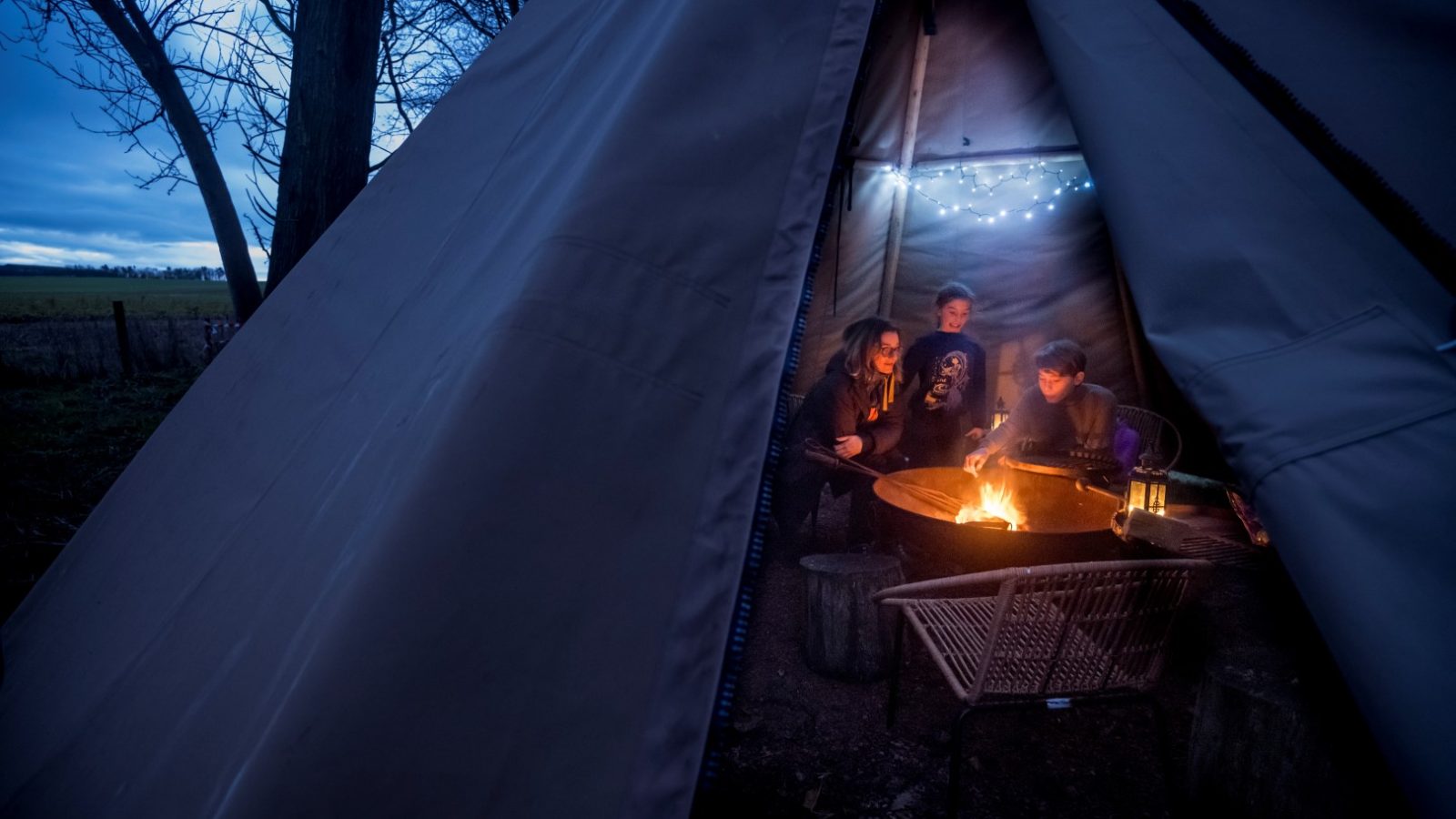 At dusk, four people gather around a campfire inside a large tent at Cambo Estate. String lights twinkle above, casting a soft glow as the trees whisper secrets of off-grid travel just outside.