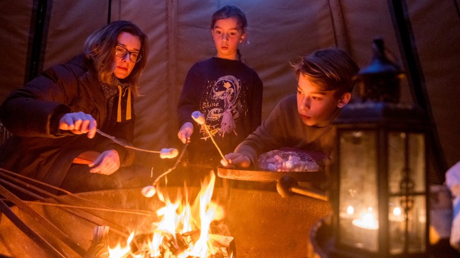 Three people roasting marshmallows over a fire inside a tent at Cambo Estate, with a lantern and string lights illuminating the cozy scene, capturing the essence of off-grid travel.