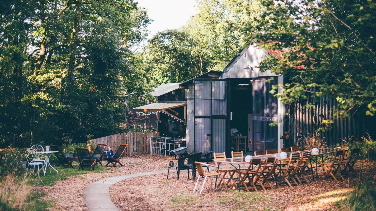 A rustic outdoor cafe surrounded by trees and greenery. Wooden tables and chairs are set up along a winding pathway. Some tables, reminiscent of Nantseren style, are covered with white tablecloths, and there's a large metal shed in the background. The scene is peaceful and inviting.