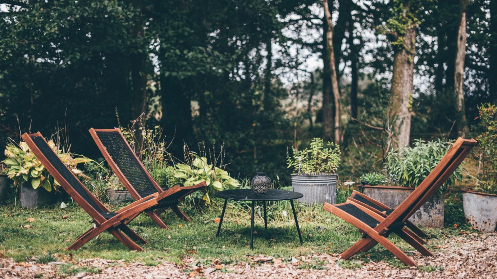 A Nantseren cozy outdoor seating area with four wooden lounge chairs around a small black metal table on a gravel surface is surrounded by potted plants and trees in the background, creating a serene and inviting atmosphere.