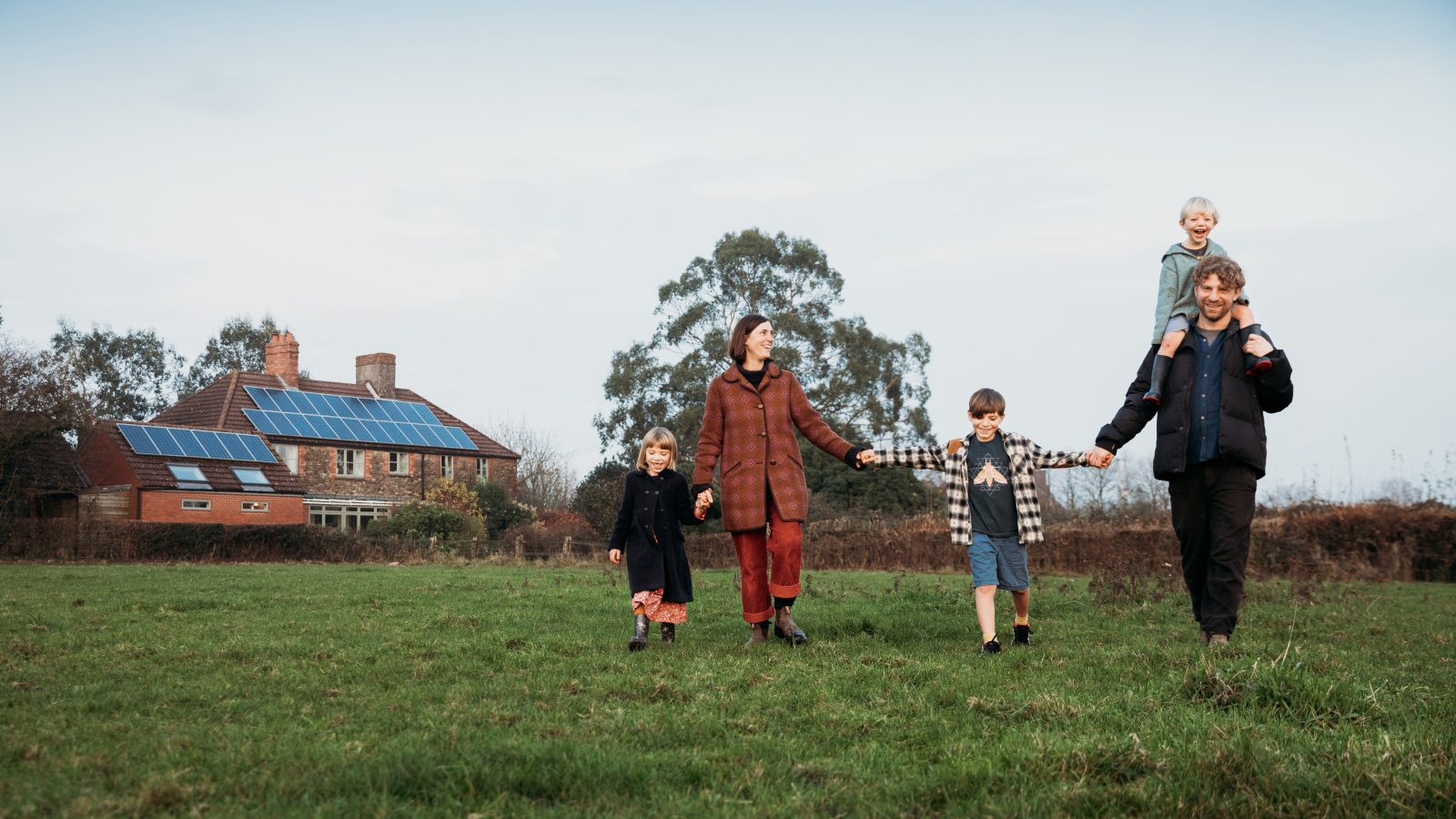 A family of five walks hand in hand across a grassy field, with a solar-paneled house and a nearby safari tent in the background.