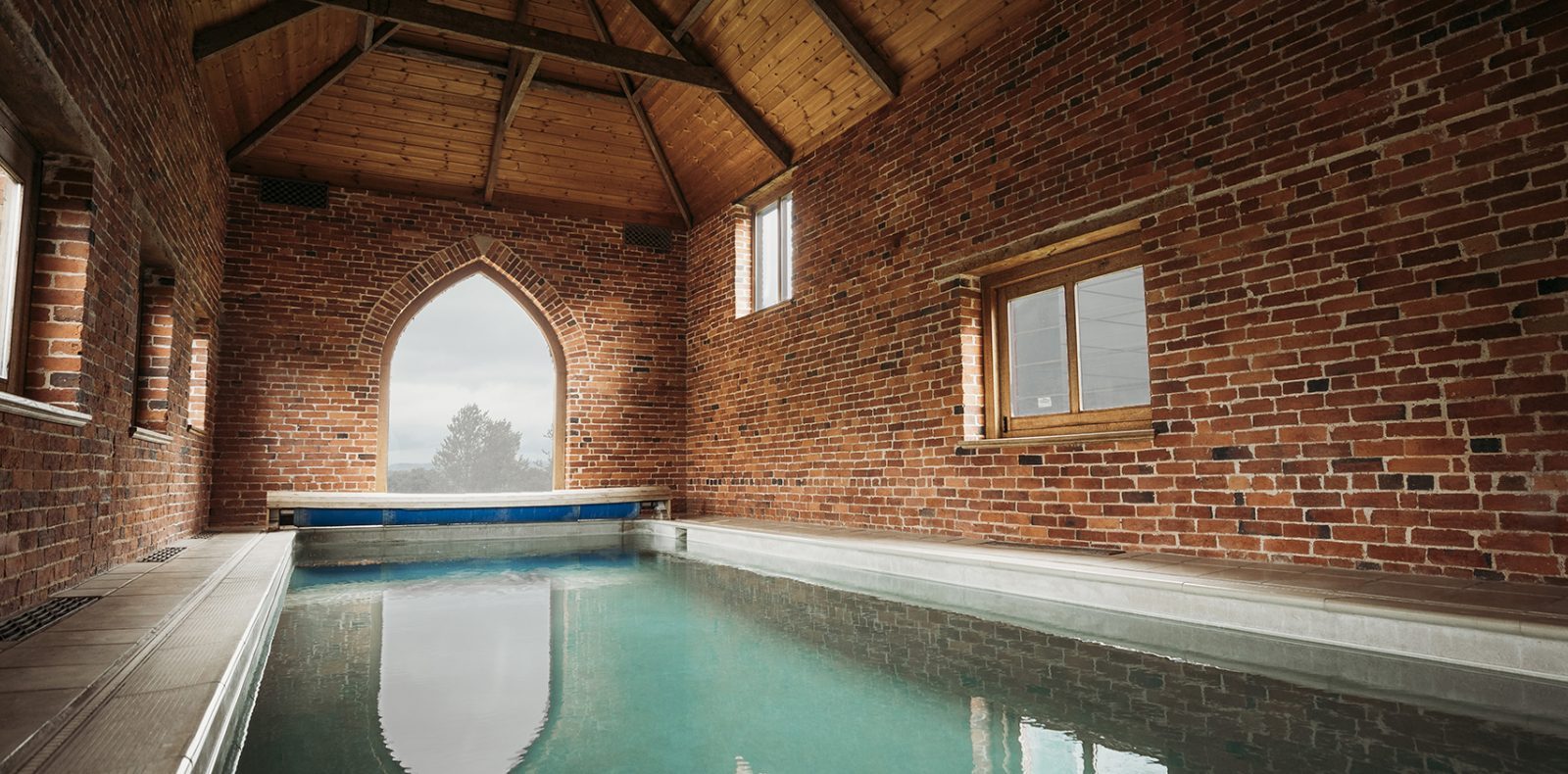 An indoor pool with calm water is housed in a brick-walled room with tall, arched windows at Pennard Farm Cottages, letting in natural light. The wooden ceiling is vaulted, and the flooring around the pool is tiled. There's a pool cover partially rolled up at one end.