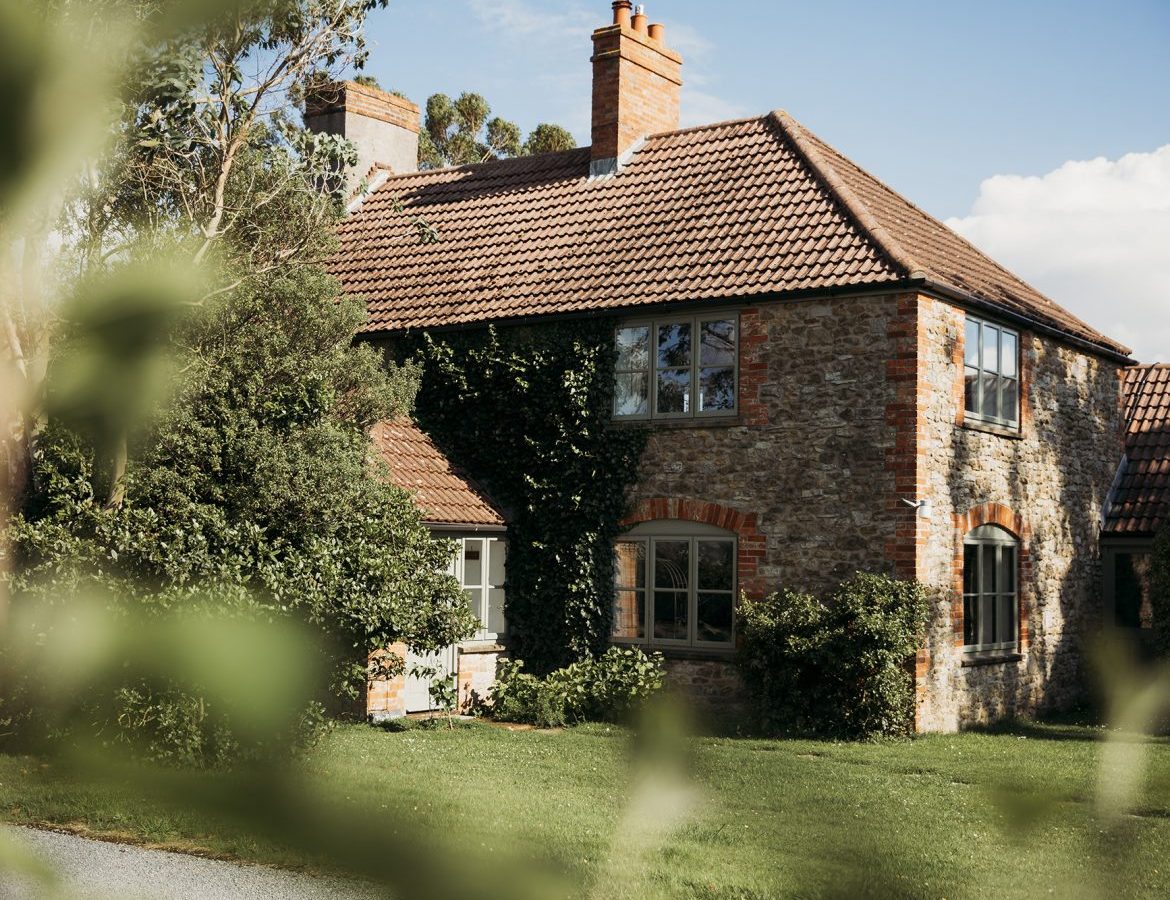 A quaint stone cottage with a tiled roof, partially obscured by greenery in the foreground, sits within Pennard Farm Cottages. The house features large windows and is surrounded by lush trees and a well-maintained lawn. The sky is clear with a few clouds.