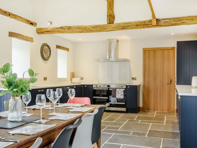 A modern kitchen and dining area with exposed wooden beams evokes the charm of a Penshenkin Barn. The kitchen features dark cabinetry, a stainless steel oven, and a vent hood. The dining table is set with placemats, wine glasses, and a vase of greenery. The floor is made of stone tiles.