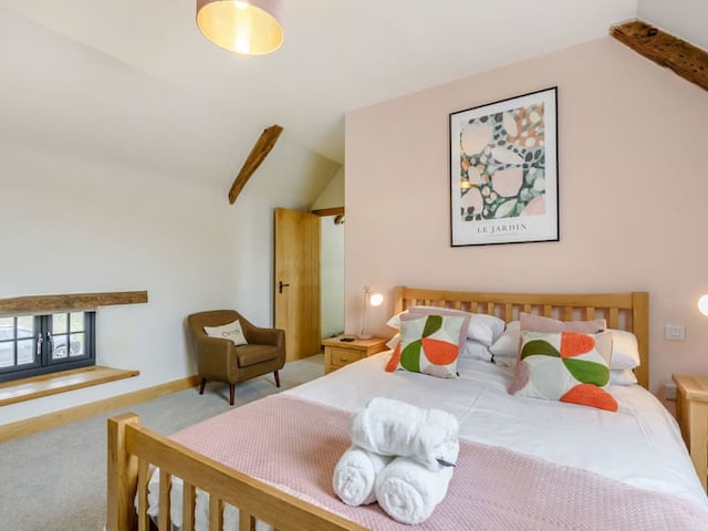 A cozy bedroom features a wooden bed with colorful pillows and a folded towel set on it. A modern armchair sits in the corner next to a small window. A framed artwork by Penshenkin hangs above the bed on the pale pink wall. Exposed wooden beams add barn-like charm to the room.