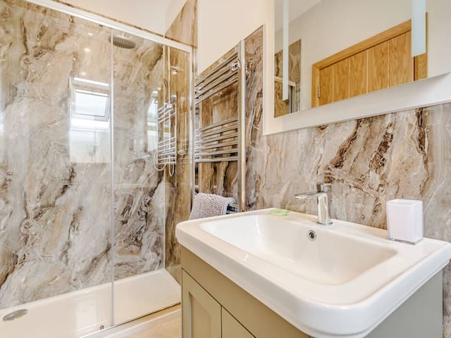 A modern bathroom featuring a large, marble-tiled walk-in shower with a glass door, a chrome towel rack, and a white rectangular sink with a faucet on a beige vanity. A mirror hangs above the sink, and a wooden barn-style door is visible in the background.