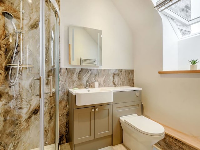 A modern barn-style bathroom with a glass-enclosed shower featuring marble-style walls, a rectangular white sink set in a grey vanity, a large mirror above the sink, a white toilet, and a small potted plant on the windowsill. Ample natural light enters through a window.