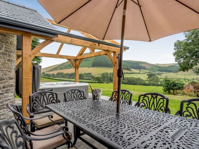 A beautifully set outdoor dining area features a cast-iron table and chairs under a large umbrella. The dining space is adjacent to the Penshenkin Barn, complete with a wooden pergola and hot tub, overlooking scenic green hills and a serene landscape in the background.