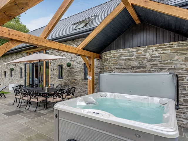 Outdoor patio featuring a hot tub with clear water, a large dining table with wrought iron chairs, and an umbrella for shade. The area, reminiscent of the Penshenkin Barn ambiance, is under a wooden pergola attached to a stone house with large windows and a slanted roof. Trees are visible in the background.