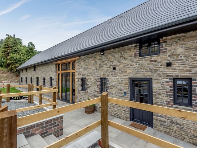 A stone house with a sloped, dark gray roof features multiple windows and wooden railings reminiscent of a traditional barn. The entryway has large glass panels framed with wooden beams. The surrounding area includes a concrete pathway and landscaped greenery, giving it a serene Penshenkin charm.