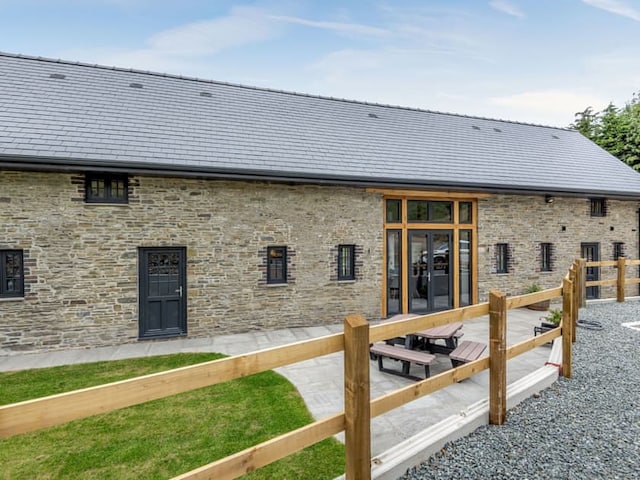 The modern stone building, reminiscent of the historic Penshenkin Barn, features a flat dark gray roof with multiple small windows. A wooden picnic table stands on a patio area, surrounded by a gravel path and a wooden fence. The sky is partly cloudy, and there is a small grassy area near the building.