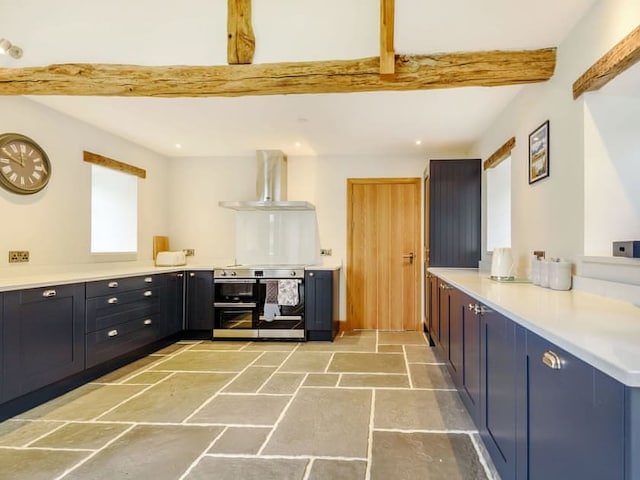 A spacious, modern kitchen featuring rustic wooden beams on the ceiling, reminiscent of a barn. It has dark blue lower cabinets with brass handles, a built-in oven, a range hood, and a stone tile floor. The left side has a clock and a window, while the right side has a door and two windows.