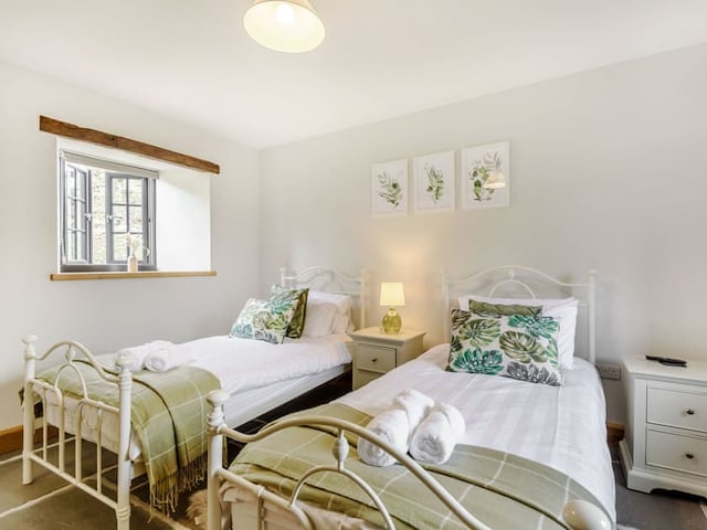 A cozy bedroom with two white metal twin beds, each adorned with green and white plaid throws and botanical print pillows. A white nightstand with a lamp is positioned between the beds. Above the beds, three botanical art prints add charm reminiscent of Penshenkin Barn. Natural light filters through a window.