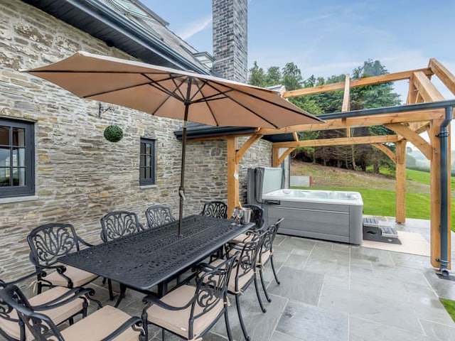 A patio with a large stone wall, featuring a black metal dining set with an umbrella. Nearby is a pergola and hot tub, and the barn adds rustic charm to the serene outdoor setting. The area is surrounded by green grass and trees.