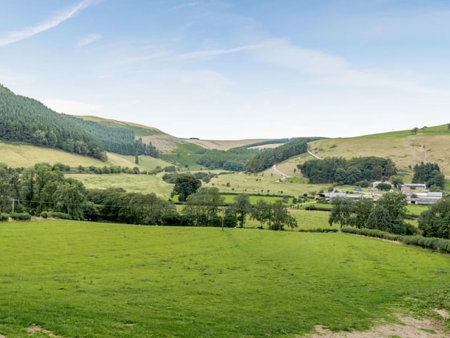 Scenic view of a lush, expansive countryside with green fields, trees, and rolling hills under a blue sky with wispy clouds. A small village or farm is visible in the distance, nestled among the greenery near the charming Penshenkin Barn.