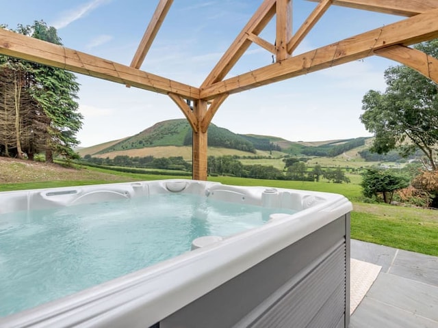 A hot tub filled with water is positioned under a wooden pergola overlooking a scenic landscape of rolling green hills and trees, with a rustic barn in the distance. The sky is clear and blue, complementing the lush greenery. The setting appears tranquil and inviting.