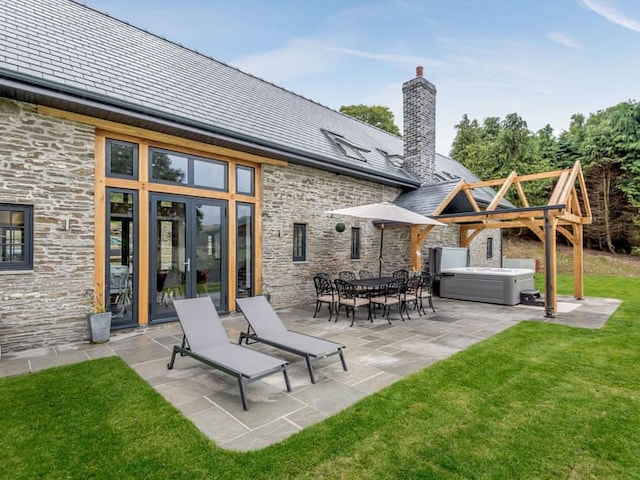 A modern stone house reminiscent of a charming barn, featuring a large, well-maintained patio with two lounge chairs, an outdoor dining table with chairs, and a covered hot tub. The home boasts large windows with wooden frames and is surrounded by a lush green lawn and trees in the background.