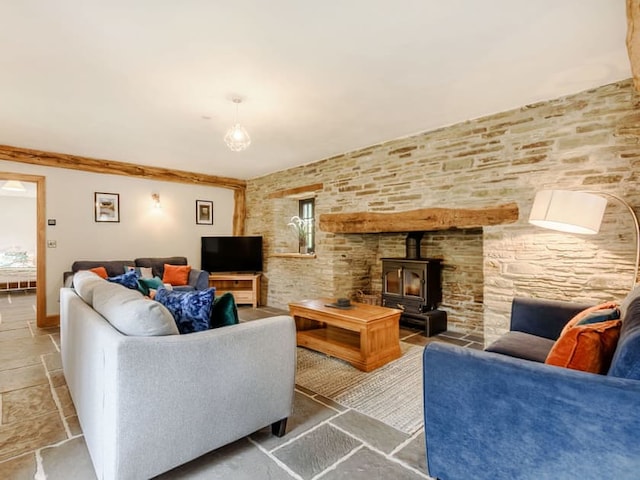 A cozy living room with stone walls evokes a barn-like charm and features a wood-burning stove framed by a wooden mantel. There's a gray sofa, blue armchair, and wooden coffee table. The space is decorated with colorful cushions, modern light fixtures, framed pictures, and a flat-screen TV.