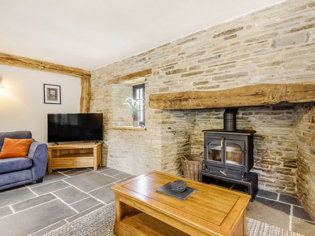 A cozy living room with stone walls and a wood-burning stove, reminiscent of the Penshenkin Barn style. A dark wooden coffee table sits in front of the stove, and a blue couch with orange cushions rests against one wall. A flat-screen TV perches on a wooden stand near a small window with a plant.