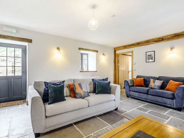 A cozy living room with two sofas: a grey one with colorful pillows and a blue one. Light-colored walls feature two windows and a wooden door reminiscent of a barn. A coffee table sits on a stone-tile floor, and the room is warmly lit by wall sconces and a hanging light.