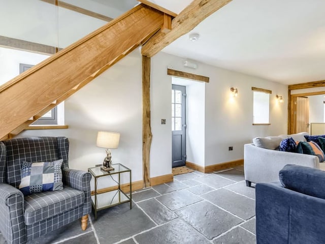 A cozy living room in the spirit of a Penshenkin Barn features a wooden staircase, stone tile flooring, and timber beams. A gray plaid armchair with a checkered pillow sits beside a small lamp on a side table. A blue sofa and a gray sofa face each other, and a door leads outside.