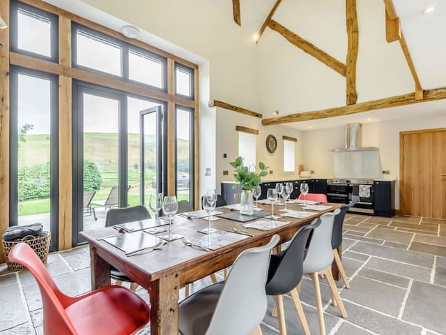 A spacious, modern dining area with a wooden table set for eight. The room, reminiscent of a barn, features high, beamed ceilings and large windows that offer a view of Penshenkin's green landscape. The kitchen area in the background has sleek appliances, including a stove and range hood.