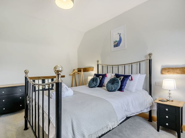 A cozy bedroom features a wrought iron bed with white and blue bedding, flanked by two wooden nightstands each bearing a lamp. The room, reminiscent of a Penshenkin barn style, is adorned with a peacock painting above the bed and has a minimalist, modern aesthetic with neutral white and gray tones.