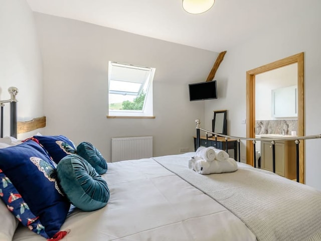 A cozy bedroom in Penshenkin Barn boasts a neatly made double bed adorned with teal and butterfly-themed pillows. The room features a slanted ceiling with a skylight, a wall-mounted TV, and an open door leading to an adjoining bathroom. Rolled towels are neatly placed on the bed.