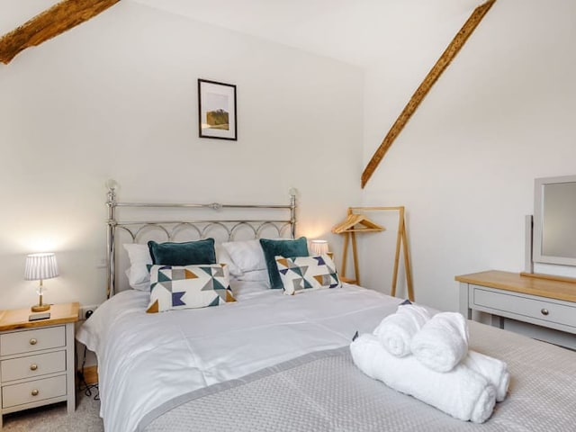 A cozy bedroom with a large bed, dressed in white linens and geometric-patterned accent pillows. The room, adorned with wooden beams reminiscent of a barn, features a framed picture above the bed, nightstands with lamps, a wooden clothing rack, and a dresser with a mirror.