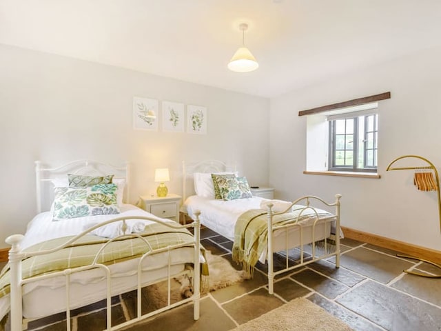 A cozy bedroom with two white metal twin beds, each adorned with green and white botanical pillows and matching throws. A small bedside table with a lamp sits between the beds. The room features a window with a countryside view of Penshenkin Barn and a modern floor lamp.