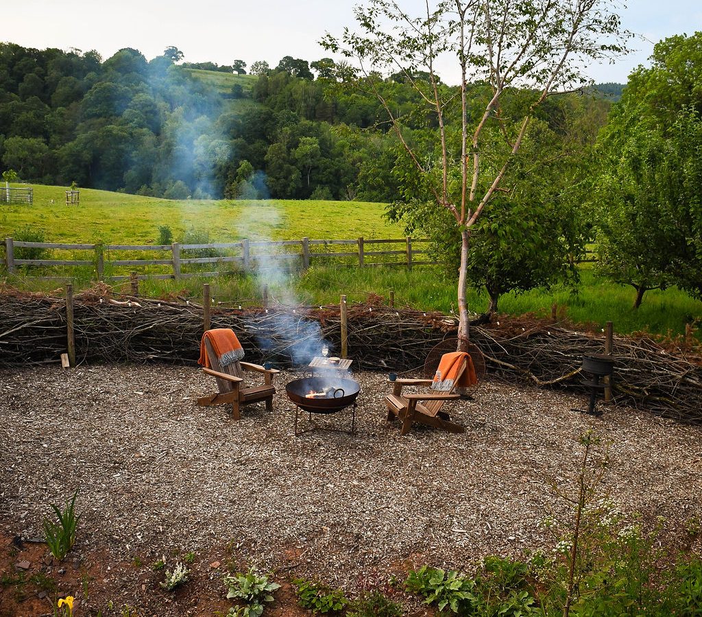 Two wooden chairs by a smoking fire pit on gravel, embraced by a wattle fence, with the lush backdrop of Perry Pond Cabin's green fields and trees.