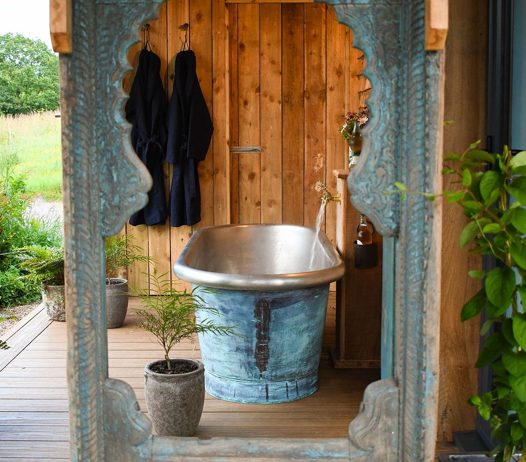 The Perry Pond Cabin boasts an outdoor wooden deck with a rustic metal bathtub set against a wooden wall. Two black robes hang to the left, and plants are elegantly arranged around the space. An intricately carved wooden doorway frames this serene setting perfectly.