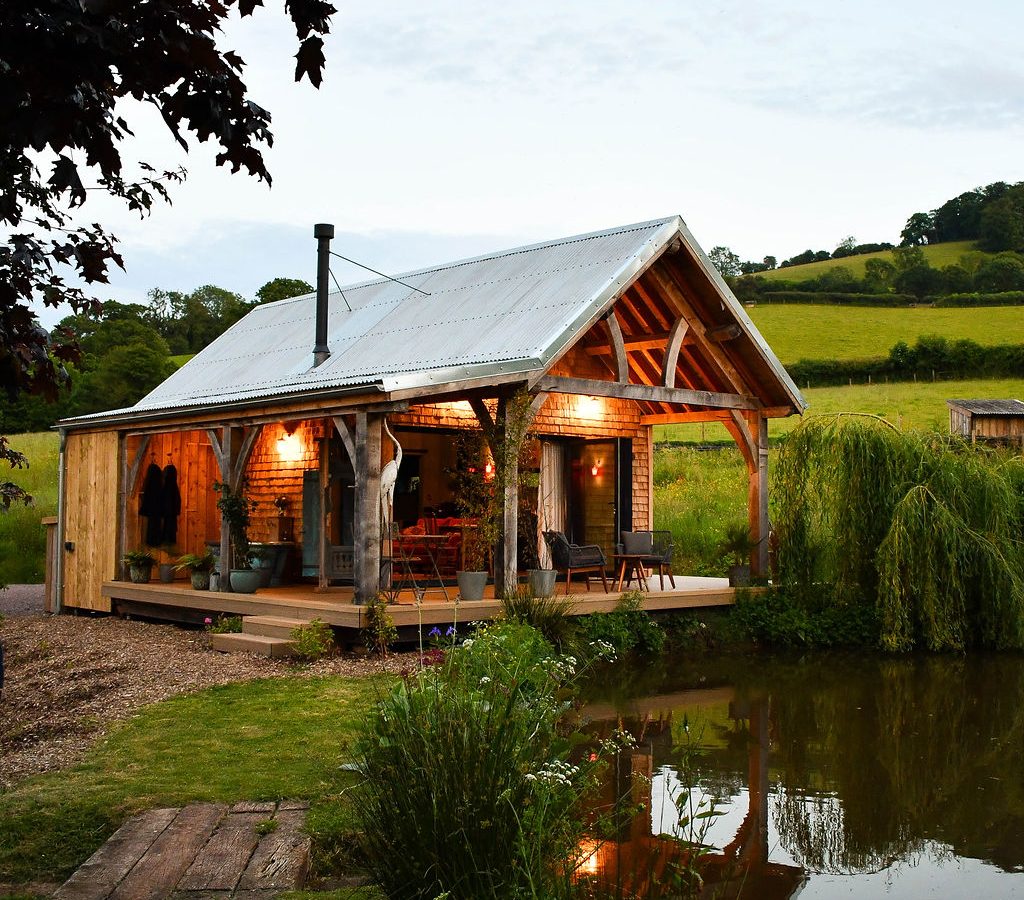 A cozy wooden cabin with a metal roof is nestled by Perry Pond. It's lit warmly, surrounded by lush greenery and rolling hills. The evening sky enhances the serene rural setting, making the Perry Pond Cabin a perfect retreat from the hustle and bustle.