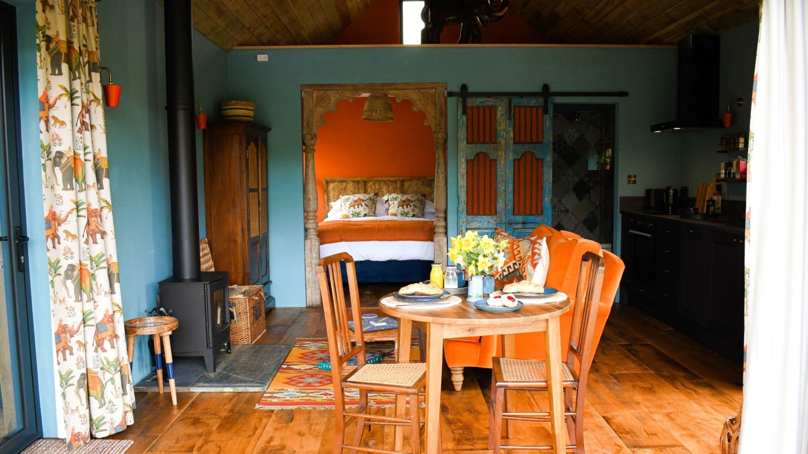 A cozy, rustic cabin interior with wooden floors, featuring a dining table set with chairs and a meal. The space includes a kitchen, a small wood stove, and a curtained bed nook with colorful bedding. Natural light pours in from large, draped windows and doors that offer views of Perry Pond.