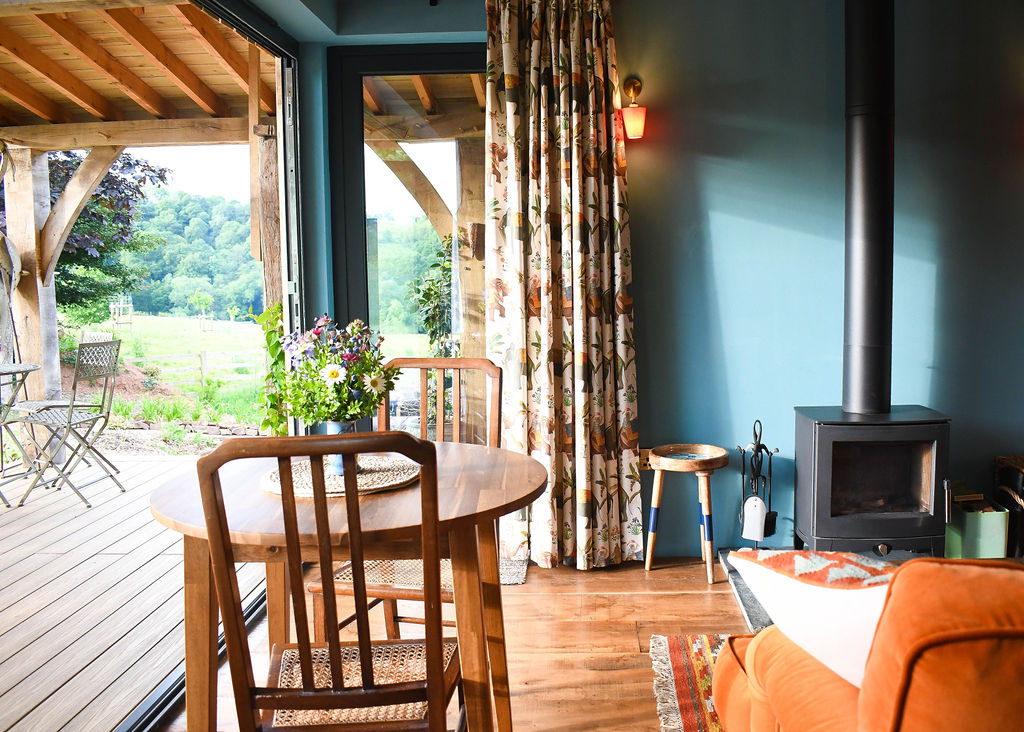 A cozy interior at Perry Pond Cabin with a round wooden table and two chairs near a sliding glass door leading to a deck with outdoor seating. The room features a wood-burning stove, floral curtains, and a vase of wildflowers on the table. Greenery and a lawn are visible outside.