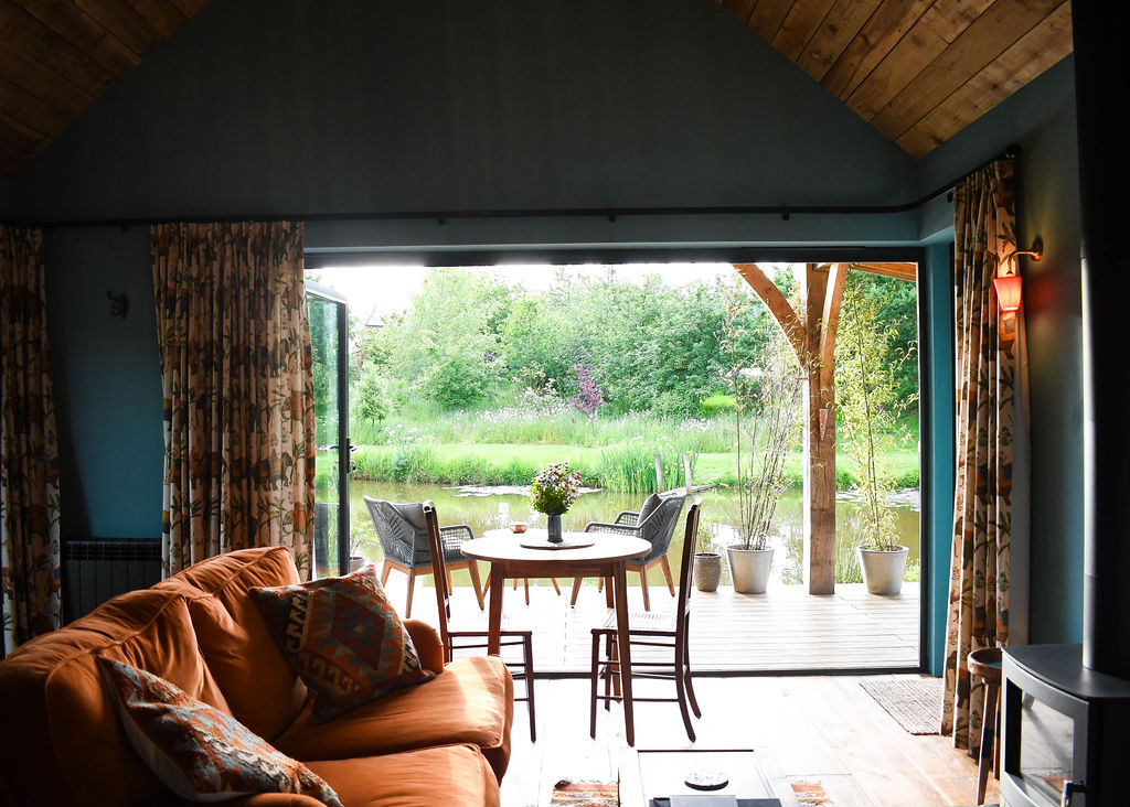 The Perry Pond Cabin interior boasts a cozy ambiance with a view of a wooden deck overlooking a serene lake and lush greenery. The room features a comfortable orange sofa, patterned curtains, a small round table with chairs, and a vase of flowers on the table. The deck has a few potted plants.