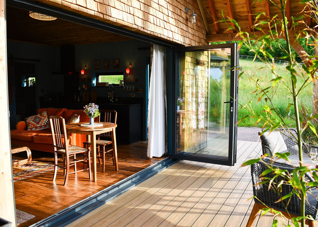 A cozy interior of the Perry Pond Cabin features a wooden floor, showcasing a dining area with a table, chairs, and a sofa. Large glass doors open to a wooden deck outside with views of greenery by Perry Pond. The ceiling is wooden, and plants adorn the inviting deck.