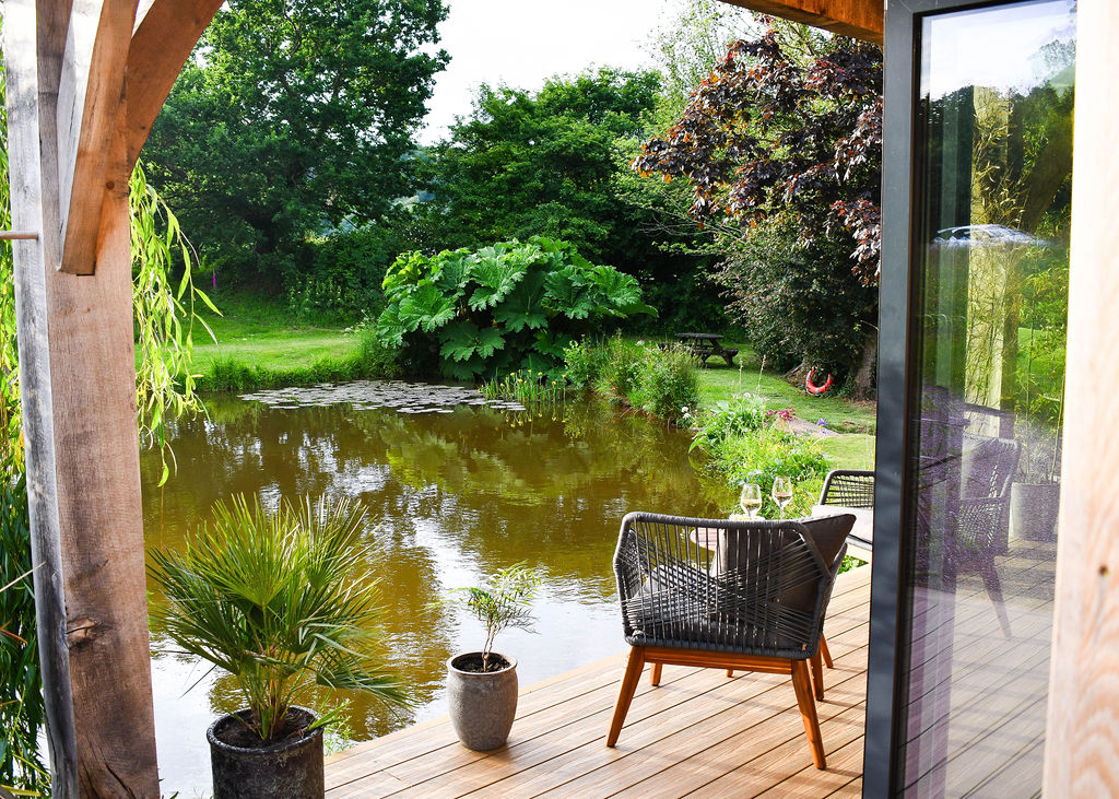 A serene wooden deck overlooking Perry Pond, surrounded by lush greenery. The deck features two black woven chairs, a small table set with wine glasses, and potted plants. It exudes tranquility with reflections of trees in the calm water and an inviting outdoor atmosphere typical of Perry Pond Cabin.